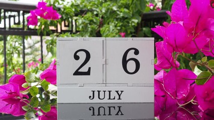 A wooden calendar with an important event for July 26 on a table with hibiscus flowers, a woman's hand sets the date on the calendar. Flip the calendar over to a cubic perpetual calendar. summer month