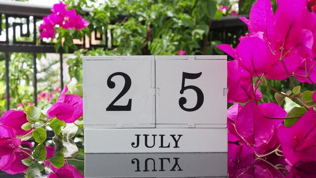 A wooden calendar with an important event for July 25 on a table with hibiscus flowers, a woman's hand sets the date on the calendar. Flip the calendar over to a cubic perpetual calendar. summer month
