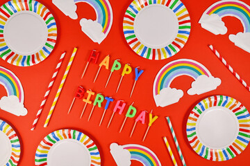 Birthday party flat lay with colorful plates, rainbow napkins, candles and drinking straws on red background