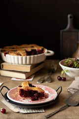 Piece of cherry pie on a vintage plate. Vintage metal kitchen utencil and retro paper on brown table. Whole shortcrust pastry pie on background. Fresh cherry in a bowl