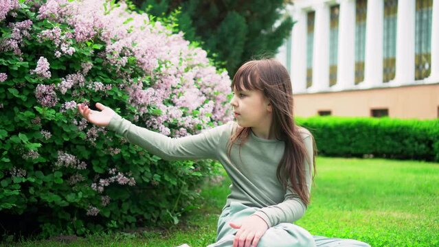 Portrait of little girl sitting on grass and touching lilac flowers on bush in park with her hand