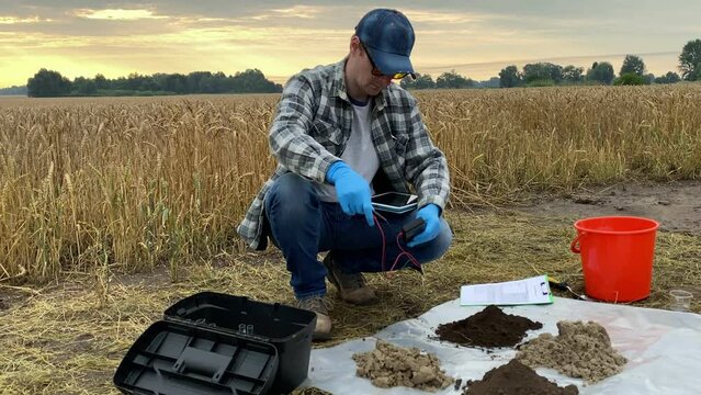 Agronomist preparing for soil measurements with digital device, sticking tester probe into sample, measuring pH temperature moisture of ground at agricultural grain field at warm summer dawn.