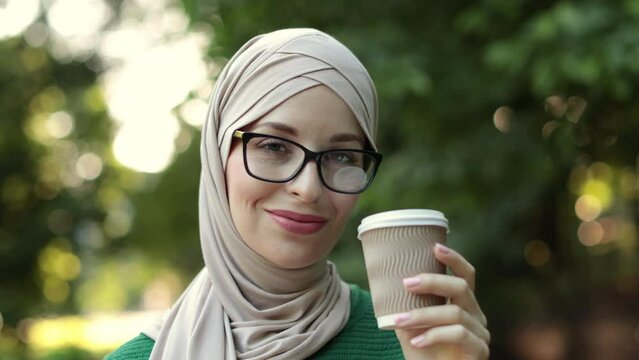 Attractive smiling muslim woman in hijab holding cup of take away coffee standing on green background of city park. Portrait of young arabian woman with eyeglass and hot drink outdoor.