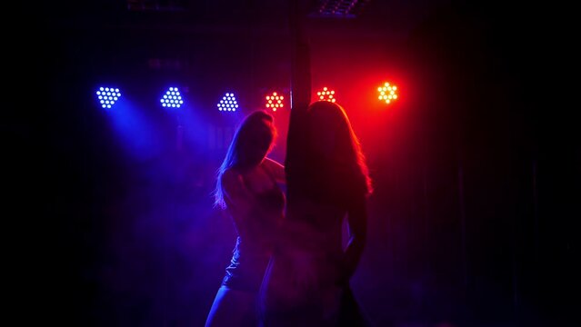 Two slender sexy women dance around a pole in the light of colorful lights with smoke in a strip club. Pole dance.