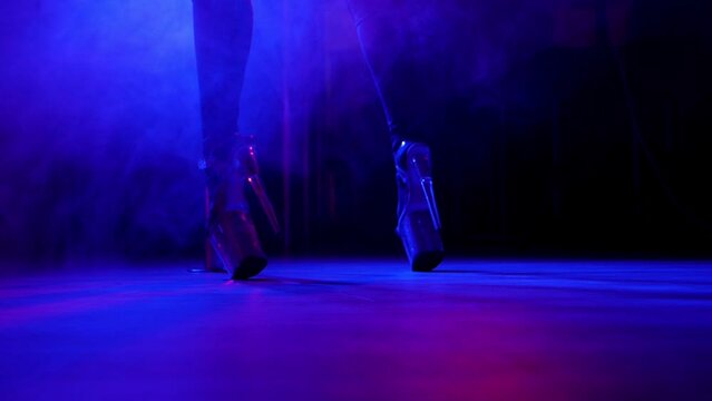 Close-up of slender legs of a dancer in high heels near a pole in a smoky nightclub under the light of multi-colored spotlights.