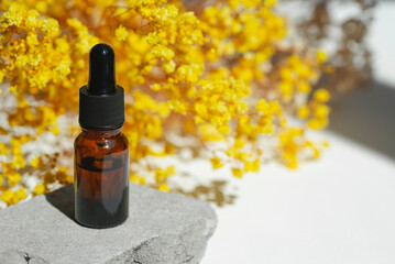 Amber dropper bottles with serum, tonic or essential oil on grey concrete podium with yellow flowers. White background with daylight. Beauty concept for face and body care