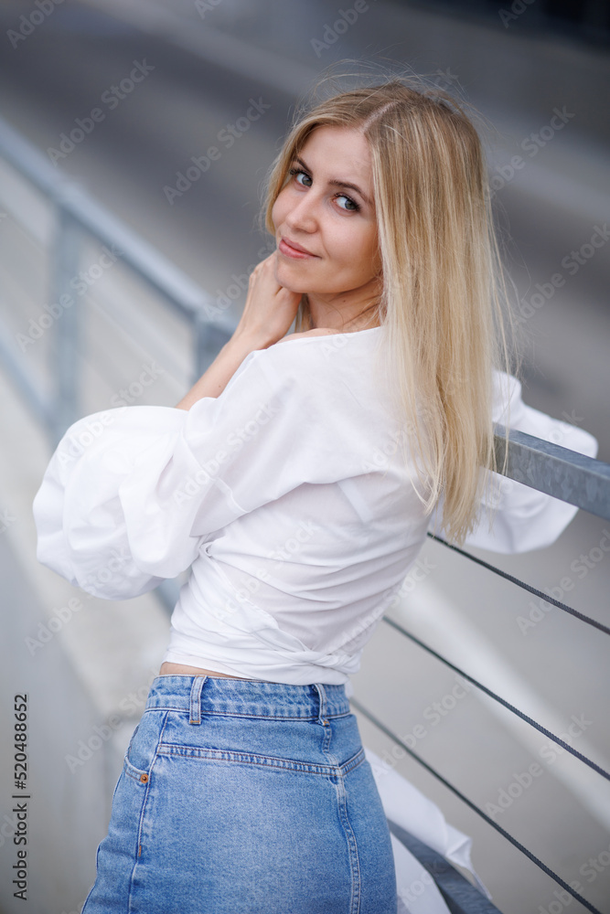 Wall mural Beautiful blonde fashionable sexy tourist girl walking in city street with long hair flying in the wind. Woman looking at camera outdoors wearing trendy white t-shirt, denim jean short skirt.