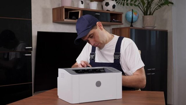 A male handyman in a blue jumpsuit repairs a printer in a client's apartment, he unscrews the screws with a screwdriver.