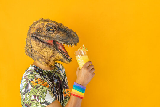 Man in dinosaur animal mask drinking cocktail wearing LGBTQ pride rainbow wristband,isolated on orange background