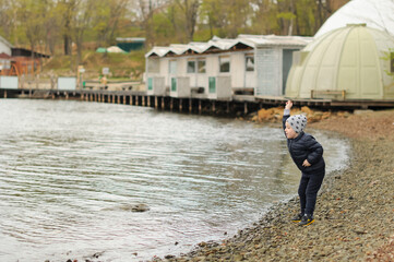 A child boy in a grey hat and dark blue jacket walks near the sea and throws stones at it. Card with selective focus