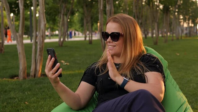 A young woman is sitting in an easy chair in the park and talking on a video call against a background of grass and trees.