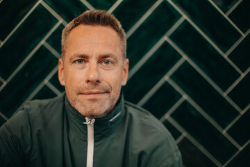 Portrait of middle-aged man in jacket sitting agiant dark tiled wall.