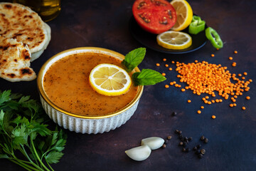 Traditional turkish lentil soup with lemon and mint in bowl, tomato and pepper on plate, flatbread, cloves of garlic and parsley on dark background. Low key photo. Selective focus. Blurred background.