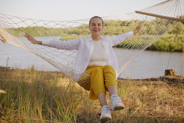 Fototapeta na wymiar Portrait of beautiful young adult woman sitting on hammock by the water, looking at camera, resting, spreads hands, laughing happily, expressing positive emotions.