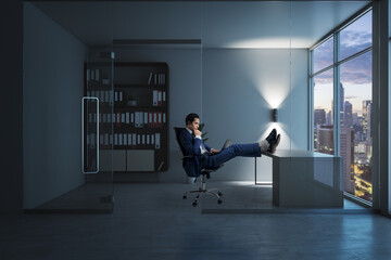 Side view of young european businessman relaxing in modern glass office interior with furniture, equipment and window with city view, with feet up on the table. CEO and executive workplace concept.