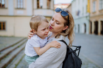 Mother consling her little daughter crying, holding her in arms in street in summer.