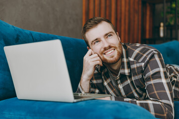 Side view young minded pensive man he wears brown shirt hold use work on laptop pc computer look aside prop up chin lying on blue sofa in own living room apartment stay home indoor flat on weekends