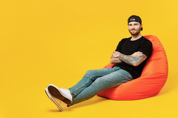 Full body young bearded tattooed man 20s he wears casual black t-shirt cap sit in bag chair hold...