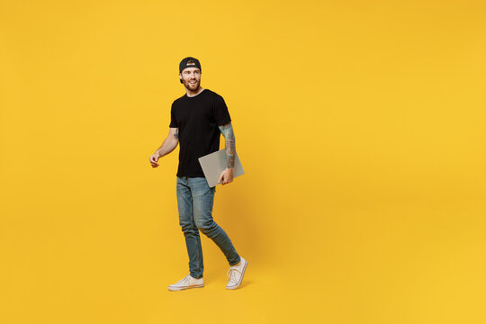 Full body young bearded tattooed man 20s he wear casual black t-shirt cap hold closed laptop pc computer look aside on workspace area isolated on plain yellow wall background People lifestyle concept - Powered by Adobe