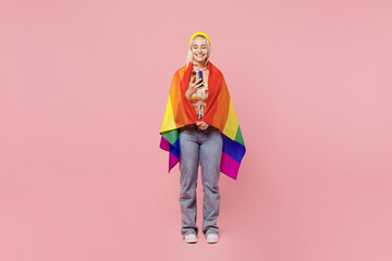 Full body young blond lesbian woman 20s wrapped in flag she wear colorful yellow hat hold in hand use mobile cell phone isolated on plain pastel light pink background. People lgbtq lifestyle concept.