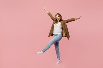 Full body young fun successful employee business woman 30s she wear casual brown classic jacket headphones listen to music raise up leg isolated on plain pastel light pink background studio portrait.