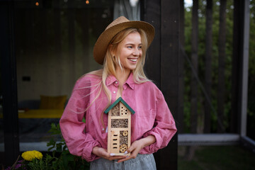 Young woman garedener holding bug and insect hotel on terrace in garden, sustainable lifestyle.