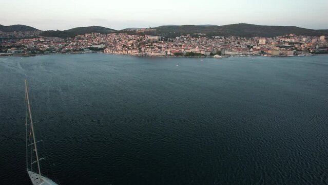 Flying towards Sibenik, old town in Croatia. Drone flight past superyacht in the Adriatic Sea. Large modern yacht anchored in front of Sibenik town. Approaching Croatian coastline on a summer evening.