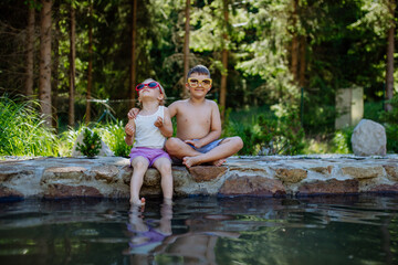 Fototapeta premium Cute girl and boy sitting together on the footpath by the lake with sun glasses, dangle their feet in the water, summer vacation concept.