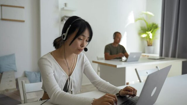 Young Asian woman with headset works in customer support advising client. Lady employee writes details of order on website via laptop closeup