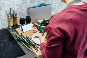 Woman tearing curly green kale leaves into pieces for kale chips or salad while watching online recipe via phone on the kitchen. Healthy eating cooking step, ready to prepare food. Selective focus.