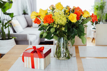 Obraz na płótnie Canvas Yellow and orange roses in a vase and gift box with red ribbon on kitchen counter table with open space living room background. Gift for Thanksgiving day, birthday, Mother's or Valentine's day.