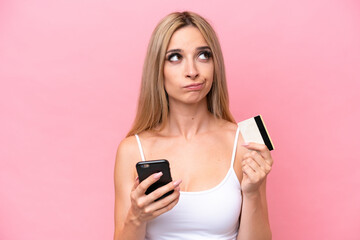 Pretty blonde woman isolated on pink background buying with the mobile with a credit card while thinking