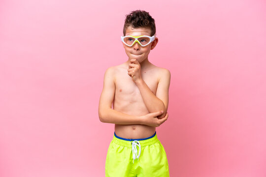 Little caucasian boy wearing a diving goggles isolated on pink background having doubts and thinking