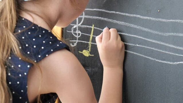 Girl, elementary school age child writing notes, musical notation on a chalkboard using a piece of chalk, blackboard drawing slow motion, closeup. Musical education, music and kids creativity concept
