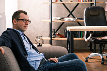 Portrait side view of Caucasian man in jacket and glasses, sitting on sofa looking away against the background of a window and shelves with documents in the office. Success and business concept.