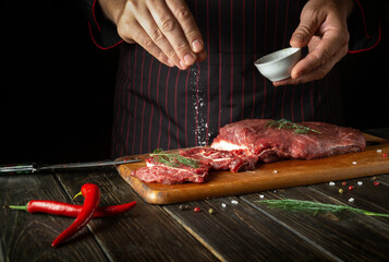 The chef salts raw fresh beef before roasting or grilling. Working environment in the kitchen of a restaurant or hotel.