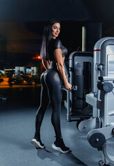 Fototapeta na wymiar Beautiful fitness brunette girl with perfect back muscles and shapes posing in the gym in a sexy black sports costume with dumbbells.