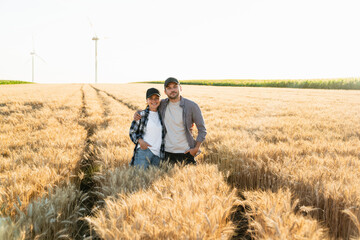 A couple of farmers in plaid shirts and caps stand embracing on agricultural field with wind turbines of wheat at sunset