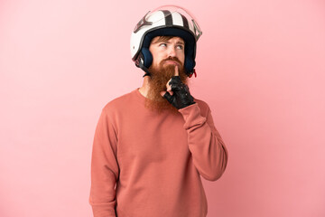 Young reddish caucasian man with a motorcycle helmet isolated on pink background having doubts while looking up