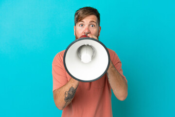 Young reddish caucasian man isolated on blue background shouting through a megaphone to announce something