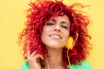 close-up of a smiling young latin woman with red afro hair wearing a blue t-shirt enjoys listening to music holding headphones with one hand on a yellow background looking at the camera