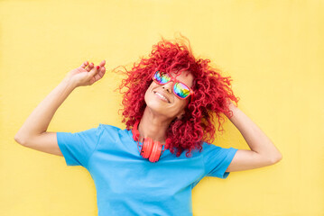 a smiling young latin woman with red afro hair wearing a blue t-shirt and sunglasses leaning on the yellow wall with her arms half raised