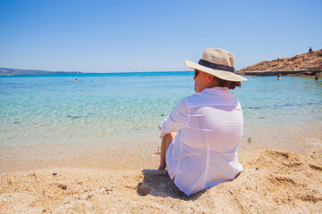 Happy woman in white shirt and hat, relaxing on sand beach, enjoying the sea view. Summer travel lifestyle, holiday concept. Beautiful seascape of turquoise sea water. Back view.	
