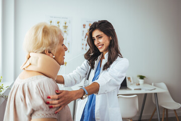 Obraz premium Female doctor putting neck orthopaedic collar on adult injured woman. Picture of senior woman having a visit at female doctor's office. Doctor helping a senior woman with her neck injury