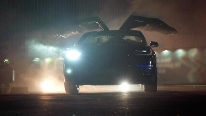 Electric car gullwing doors raising up with headlights flashing in darkness. Wide shot modern contemporary automobile at dark night outdoors. Luxury and auto industry