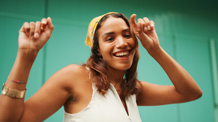 Cute tanned woman with long brown hair wearing white top and yellow bandana listens to music in wireless headphones and dances on blue wall background.