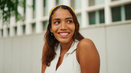 Smiling cute tanned woman with long brown hair wearing white top and yellow bandana seated on bench on cityscape background