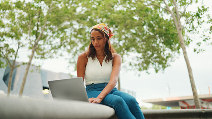 Cute tanned woman with long brown hair in white top and yellow bandana using laptop while sitting...
