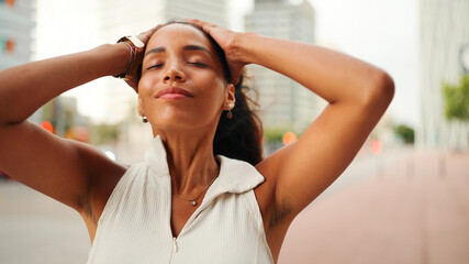 Smiling cute tanned woman with long brown hair wearing white top raises her hands and touches her hair. The girl enjoys the breeze