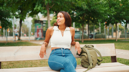 Lovely tanned woman with long brown hair wearing white top sitting relaxed on park bench looking away. Life style concept. Slow motion.